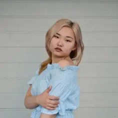 Młoda kobieta w niebieskiej sukience w paski, z blond włosami, na tle białej ściany.